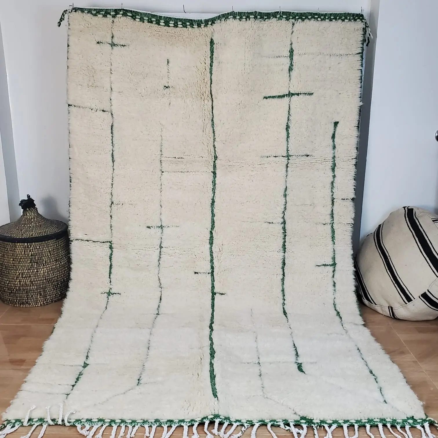 De 100% HANDGEMAAKT MAROKKAANS TAPIJT 150 - 250 cm nr 51, een wit tapijt met groene verticale en horizontale lijnen, ligt uitgespreid op een houten vloer. Ernaast liggen een gevlochten mand en een gestreept kussen. Het tapijt heeft ook franjes aan de rand.