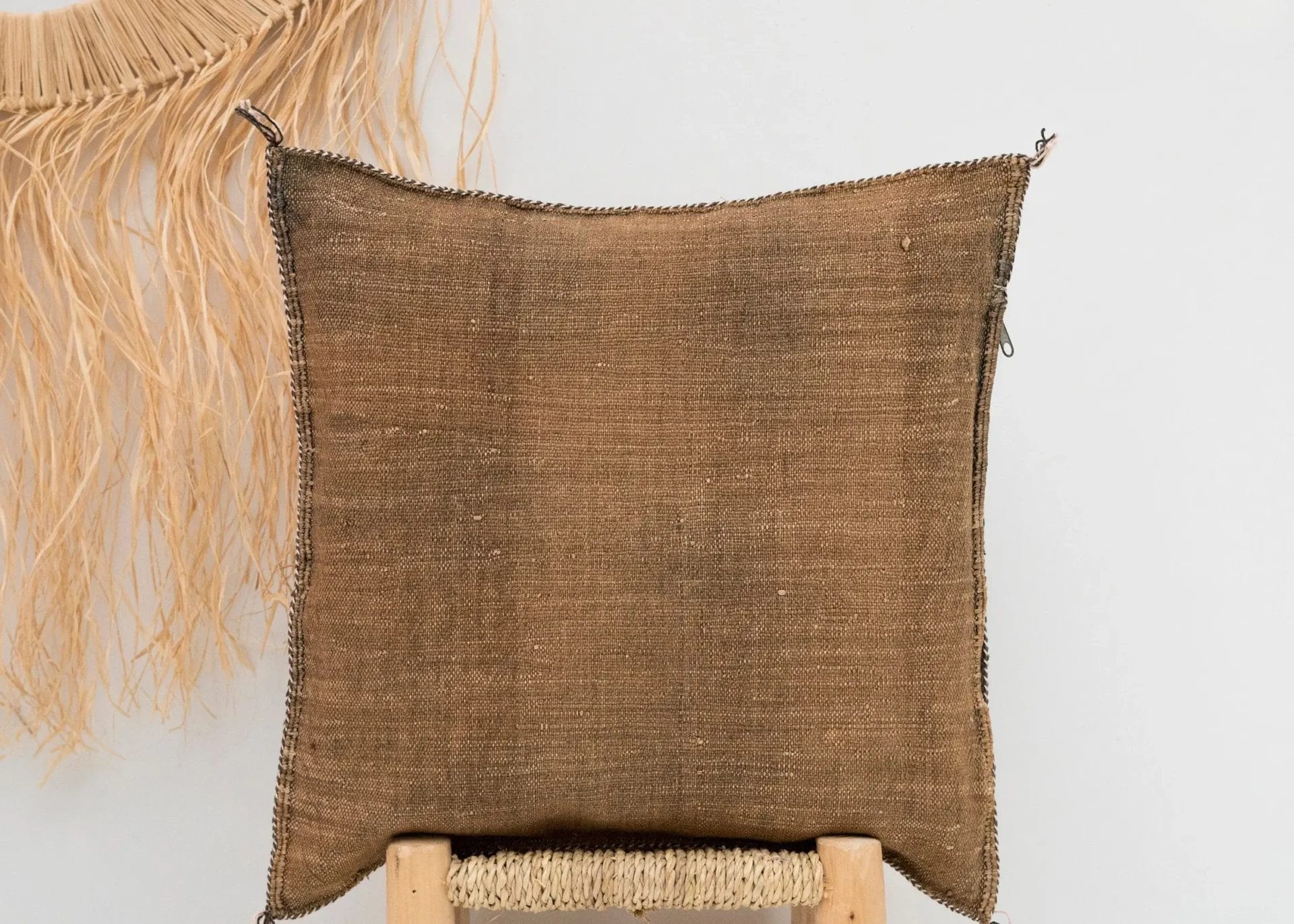 Een vierkant, bruin kussen met textuur op een lichtgekleurde houten kruk. Een decoratieve natuurlijke vezelfranje hangt aan de muur op de achtergrond.