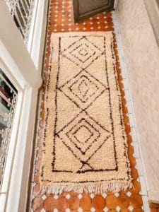 Een beige tapijt met een geometrisch ruitpatroon ligt op een betegelde vloer in een smal pad buiten met een ijzeren leuning aan de zijkant.