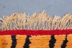 Franje van een kleurrijk geweven tapijt met rode, oranje en zwarte strepen op een grijs oppervlak.