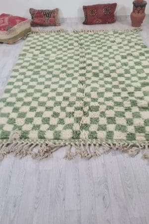 Een groen en wit 250 - 150 cm Beni Ouarain vloerkleed 2136 Marokkaans, handgemaakt met franje randen, ligt op een lichte houten vloer. De achtergrond bestaat uit decoratieve kussens en een kleine potplant.