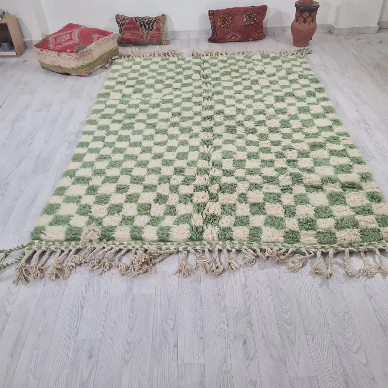 Een groen en wit 250 - 150 cm Beni Ouarain vloerkleed 2136 Marokkaans, handgemaakt met franje randen, ligt op een lichte houten vloer. De achtergrond bestaat uit decoratieve kussens en een kleine potplant.