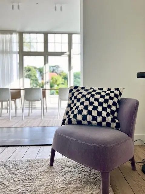 Een paarse stoel met een zwart-wit geblokt kussen staat op een licht tapijt in een lichte kamer met grote ramen en uitzicht op een eettafel met stoelen.