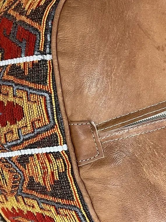 Close-up van een bruine leren tas met een kleurrijk, ingewikkeld kralenpatroon, met rode, gele, zwarte en witte kleuren. Een zichtbare naad met stiksels loopt langs de rand.