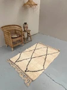 Een rieten stoel en een klein houten tafeltje met een vaas en bord staan naast een beige Baby Beni Ouarain-tapijt van 165x101 cm met zwarte kruispatronen op een grijze vloer.