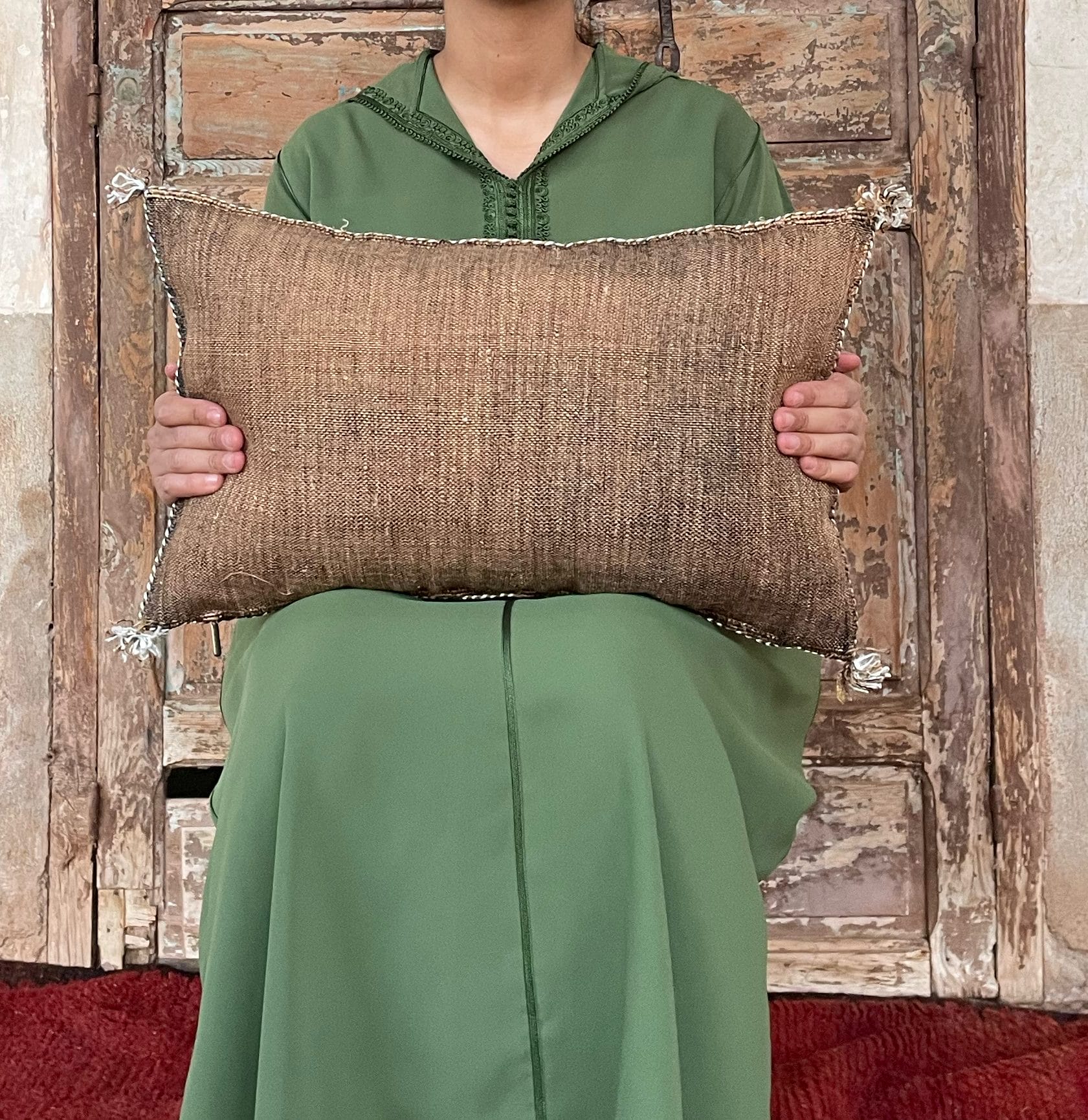 Een persoon in een groen gewaad zit met een bruin geweven kussen in zijn hand tegen een rustieke houten achtergrond.