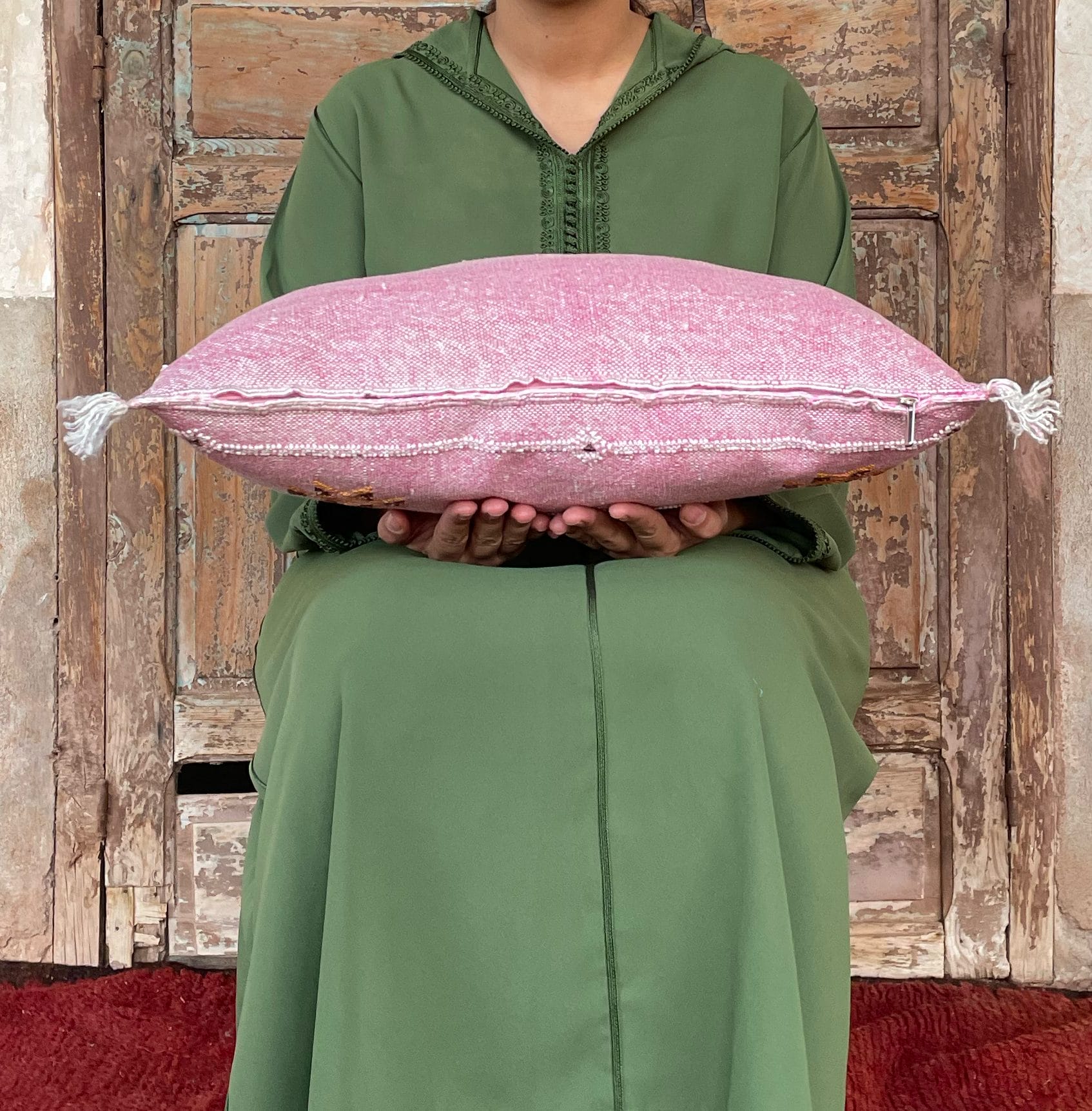 Persoon in het groen met een roze kussen met kwastjes, zittend voor een rustieke houten deur.