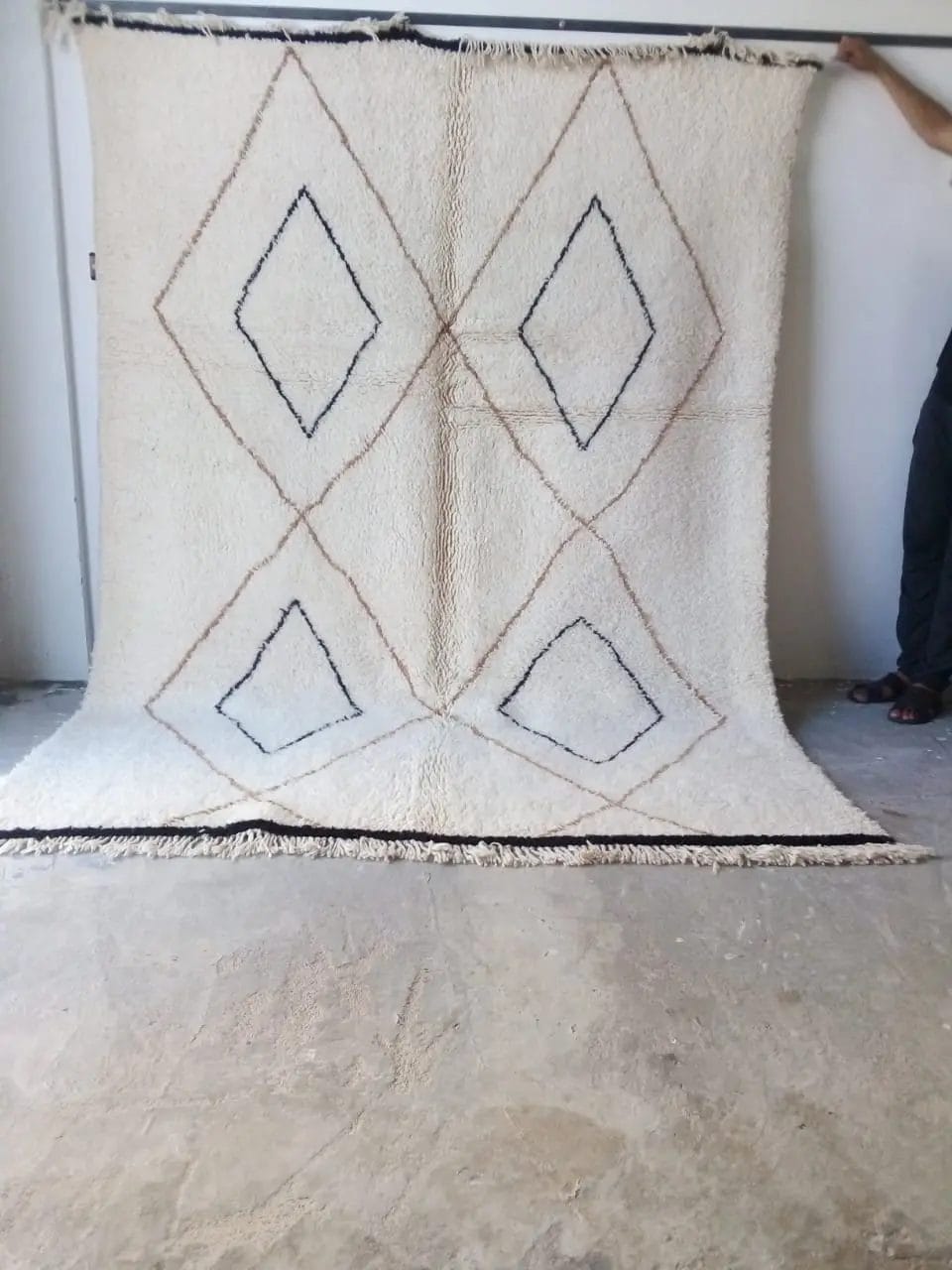 A person holds up a large 54 HANDMADE MOROCCAN BENI OUARAIN RUGS, decorated with a geometric diamond pattern in brown and black lines, displayed against a plain, light-colored wall.