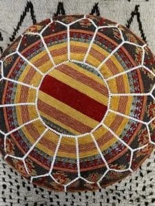 Bovenaanzicht van een kleurrijk geweven rond kussen met radiale strepen en een wit netpatroon op een grijs oppervlak.
