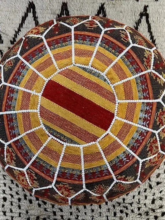 Bovenaanzicht van een kleurrijk geweven rond kussen met radiale strepen en een wit netpatroon op een grijs oppervlak.