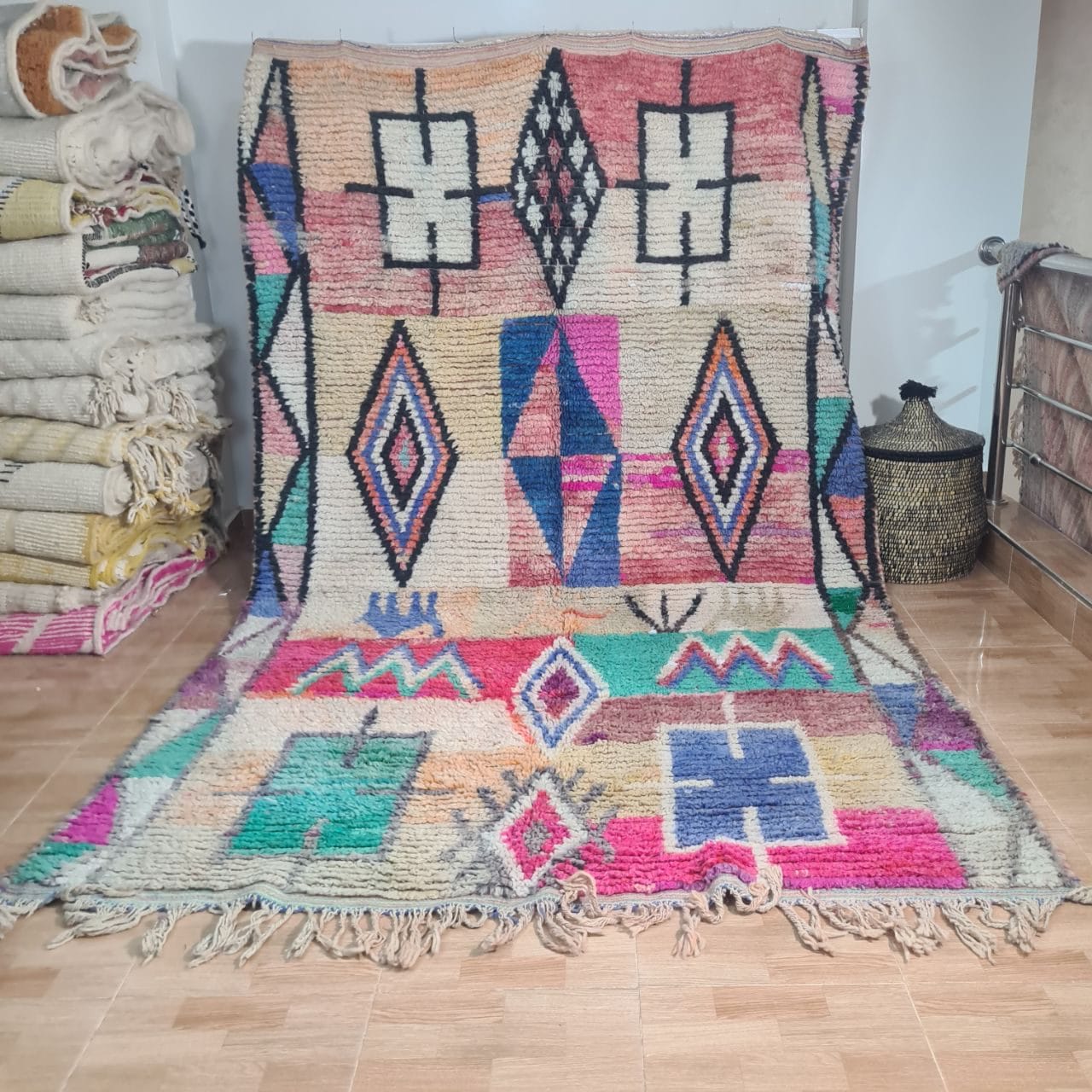 Een HANDGEMAAKT MAROKKAANS TAPIJT Boujaad 200 x 300 cm met levendige geometrische patronen en franjes staat op een hardhouten vloer, met opgevouwen dekens en een gevlochten mand op de achtergrond.