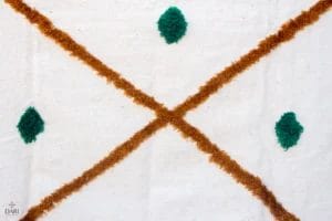 Een gestructureerde stof met een bruin "X"-patroon dat groene ruitvormen kruist op een crèmekleurige achtergrond.