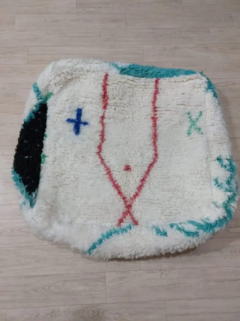 Een MAROKKAANSE HANDGEMAAKTE BENI OUARAIN BERBER POEF 004 met een vierkante vorm, een textuurontwerp en geometrische patronen in rood, blauw, groen en zwart op een lichte achtergrond is op een houten vloer geplaatst.