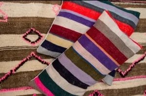 Twee levendige Kilim Vintage kussens - kleurrijk zijn geplaatst op een patroon vloerkleed met ruit- en lijnontwerpen. De kussens tonen horizontale strepen in een spectrum van kleuren zoals rood, paars, roze en groen.