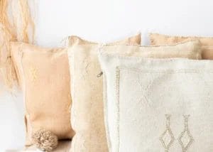 Beige en lichtbruine kussens met eenvoudige gestikte patronen zijn op een witte achtergrond gestapeld.