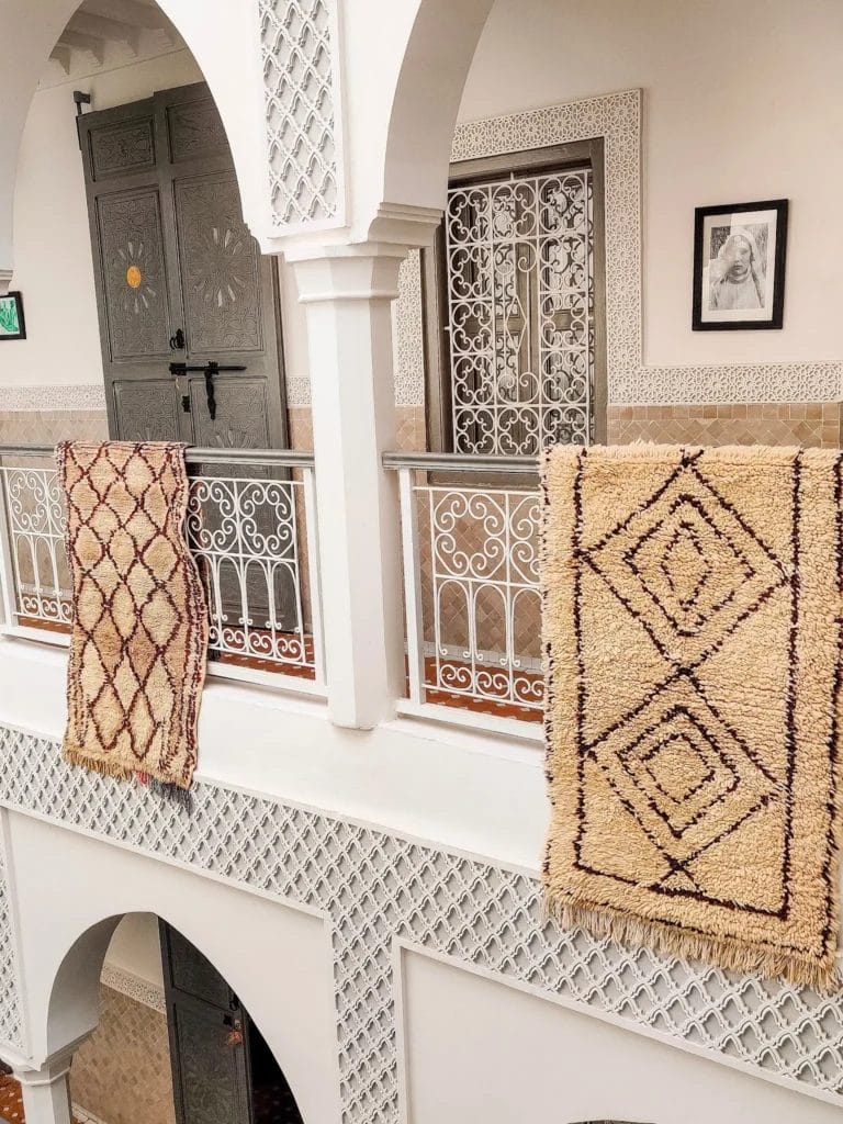 Twee Azilal vloerkleden met bruine ruitpatronen hangen over een witte balustrade op de bovenste verdieping van een rijkversierd gebouw met ingewikkeld traliewerk en ingelijste schilderijen aan de muren.