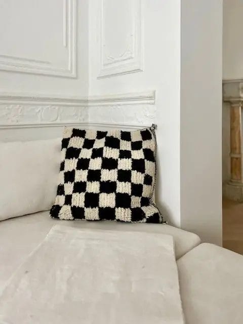 Zwart-wit geblokt kussen op een witte bank in een minimalistische kamer met witte muren.