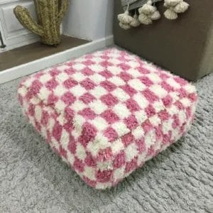 Een Marokkaanse Berber poef, BABY PINK, met een ruitjespatroon staat op een grijs tapijt in een kamer met een cactus en met kwastjes versierde meubels op de achtergrond.