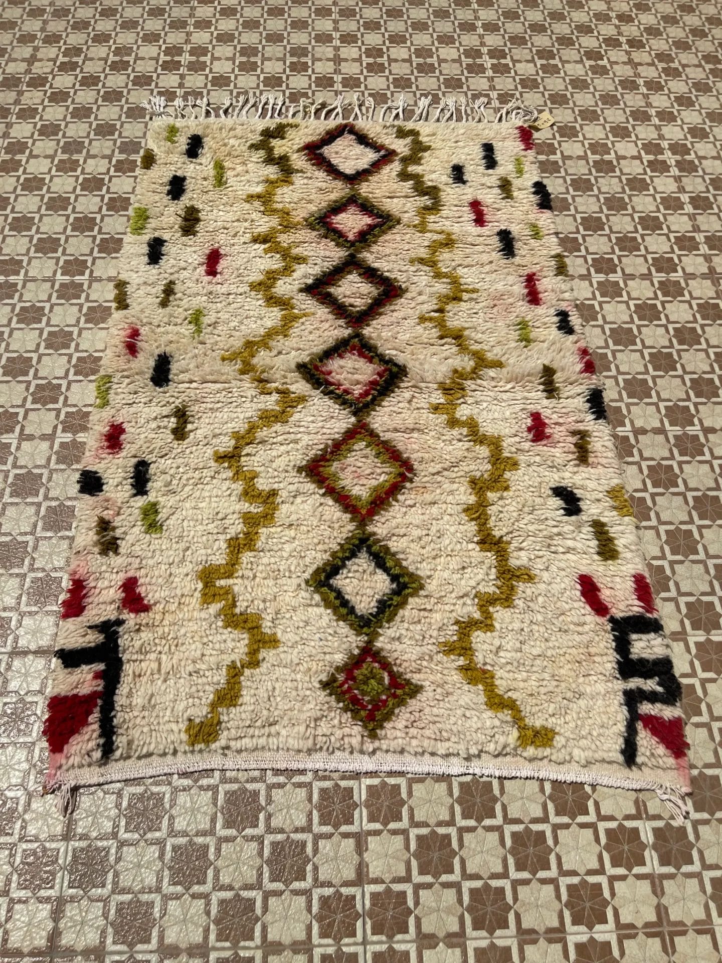 Een decoratief tapijt met geometrische patronen in rood, zwart, geel en groen op een crèmekleurige achtergrond, gelegd op een betegelde vloer met een vierkant ontwerp.