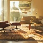 De zonnige woonkamer is voorzien van een leren loungestoel, een voetenbank, een beige bank en een salontafel op een vloerkleed met patroon en vloerverwarming. Alles is voorzien van een groot raam met uitzicht op het weelderige groen.