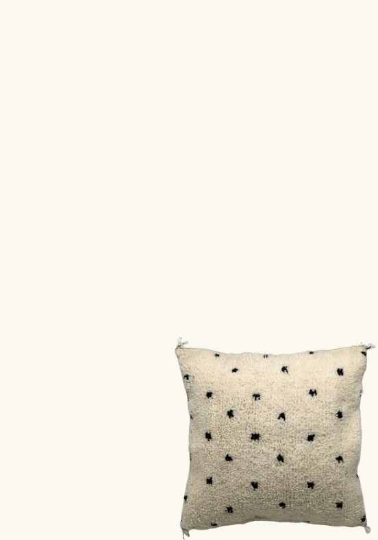 Een vierkant Kussens-kussen met een textuur, een crèmekleurige achtergrond en een klein zwart stippenpatroon, wordt afgebeeld op een effen lichte achtergrond. Het lijkt op een subtiel maar elegant item dat je op een exclusief designmenu zou kunnen vinden.