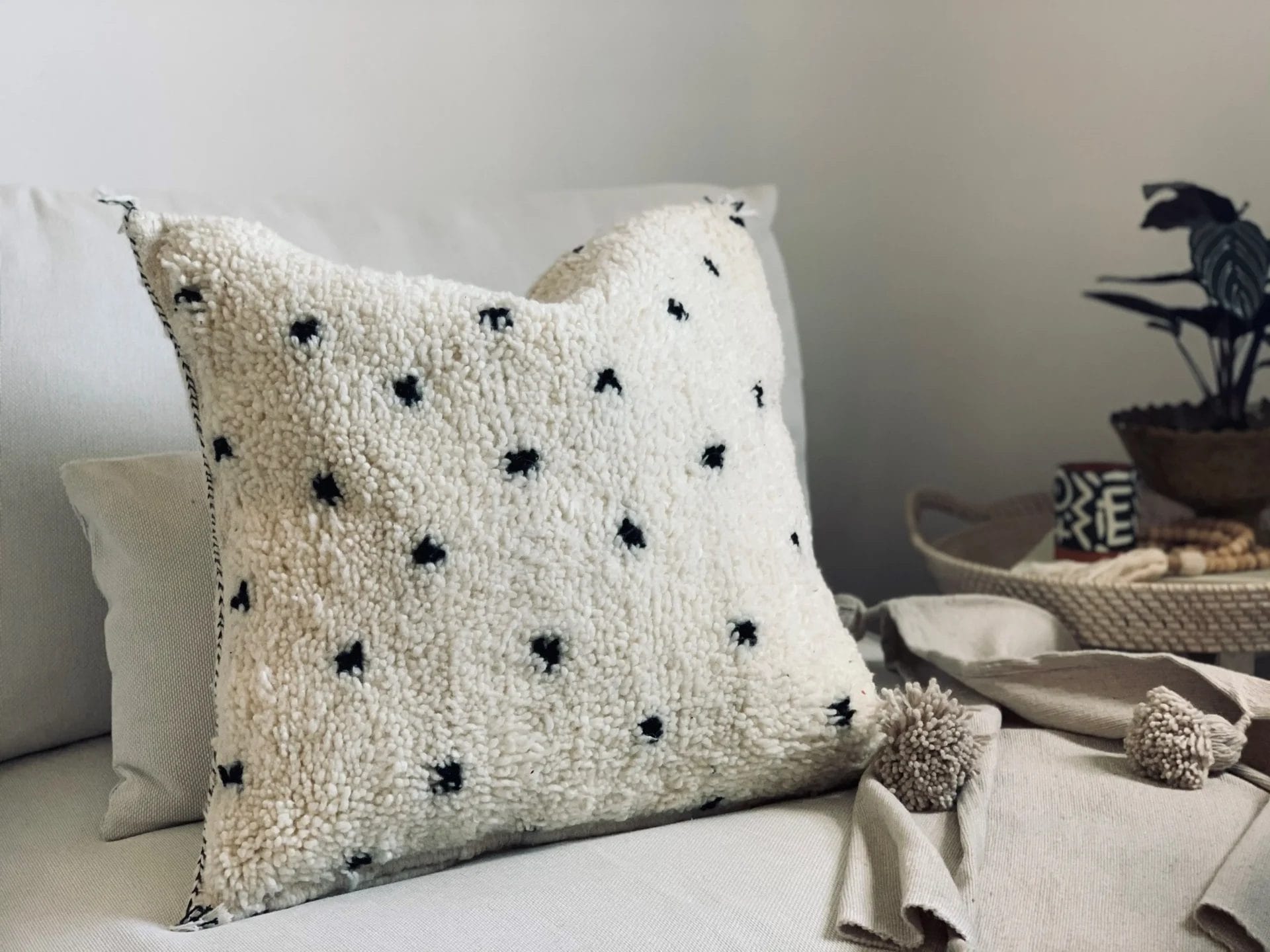 Een zacht wit kussen versierd met zwarte stippen ligt op een lichtgekleurde bank van Houseify, samen met een plaid met textuur en een gevlochten mand met een plant en decoratieve voorwerpen.