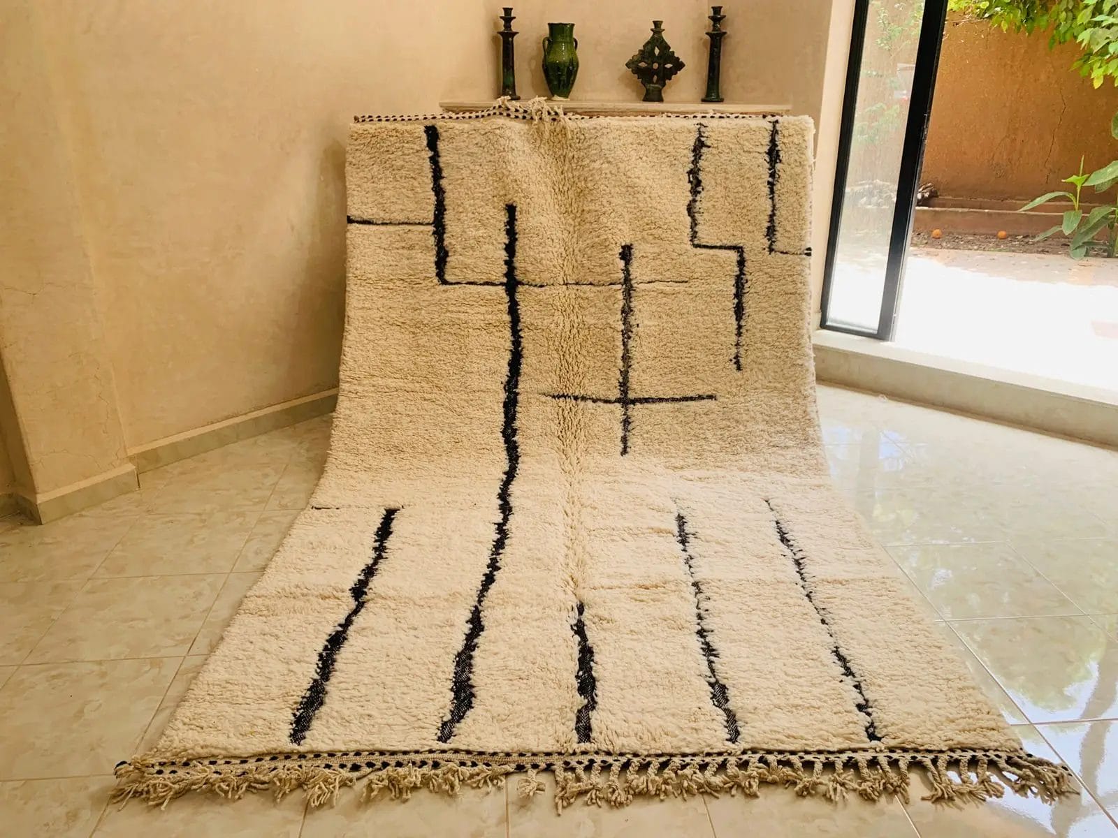 Het BENI OUARAIN VLOERKLEED 2065 MAROKKAANS TAPIJT HANDGEMAAKT 185 - 285 cm, met zwarte geometrische lijnen en franje aan de randen, staat op een witte tegelvloer bij een raam, tegen een muur die versierd is met decoratieve voorwerpen op een plank erboven.