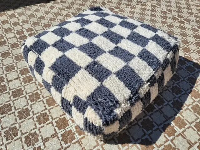 Een vierkant, zacht kussen met een zwart-wit geblokt patroon ligt op een tegelvloer.