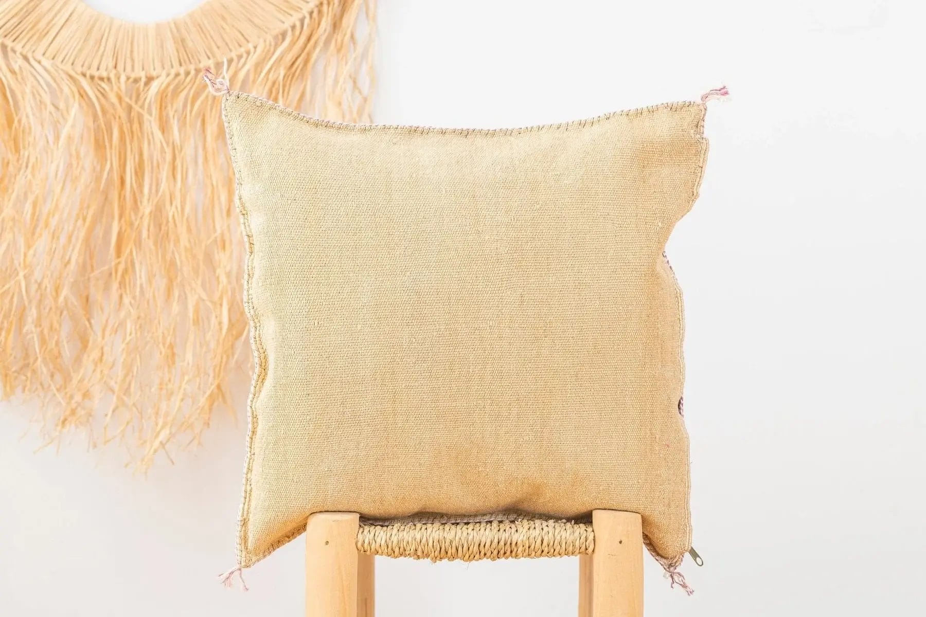 Een beige vierkant kussen ligt op een houten kruk met een gevlochten strodecoratie op de achtergrond tegen een witte muur.