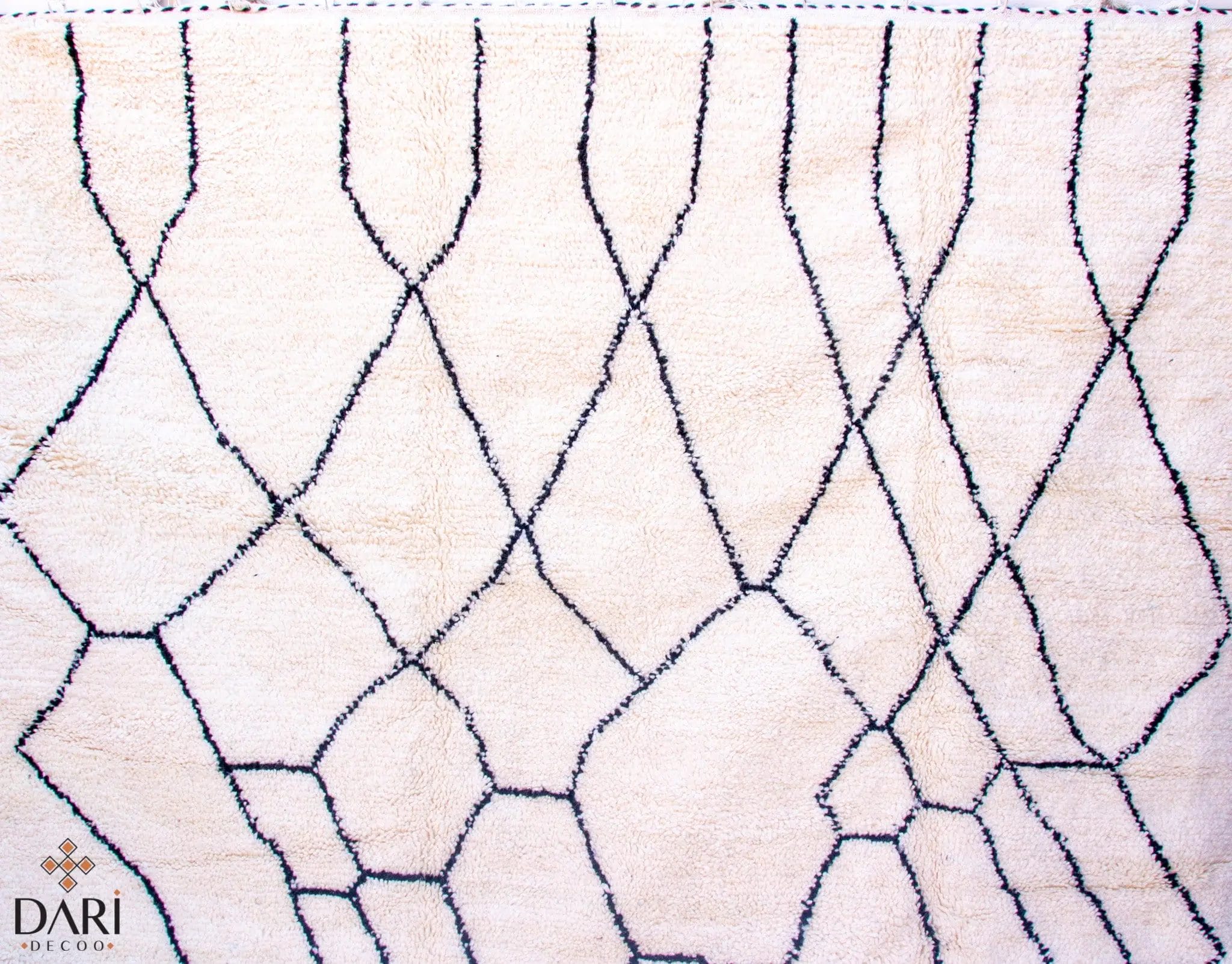 Een crèmekleurig tapijt met een onregelmatig zwart lijnenpatroon dat abstracte vormen creëert. Logo in de linkerbenedenhoek.