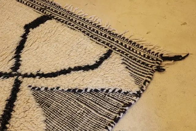 Een hoek van een wit en zwart kleed met franjes ligt op een beige vloer.