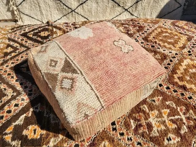 Een vierkant kussen met geometrische patronen en een mix van beige, roze en bruine kleuren, geplaatst op een beige en bruine Kleurrijke poef met vergelijkbaar patroon.