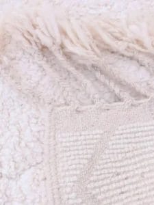 Close-up van een geweven, wit textiel met textuur, franjes en een geometrisch reliëfontwerp.