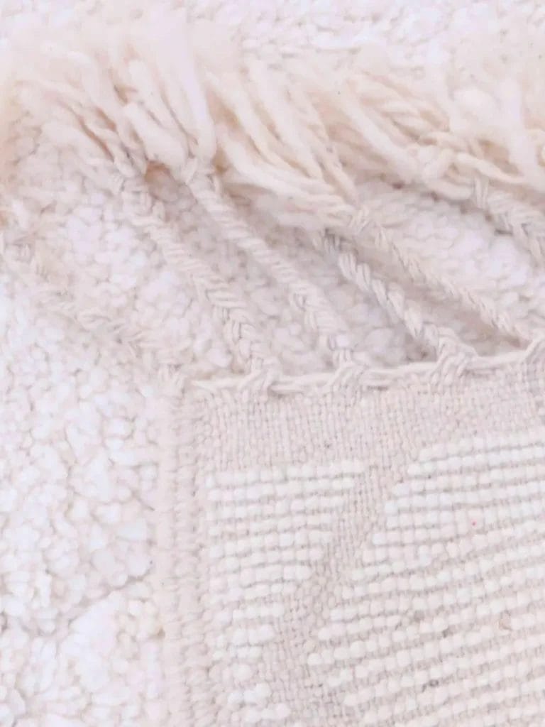 Close-up van een geweven, wit textiel met textuur, franjes en een geometrisch reliëfontwerp.