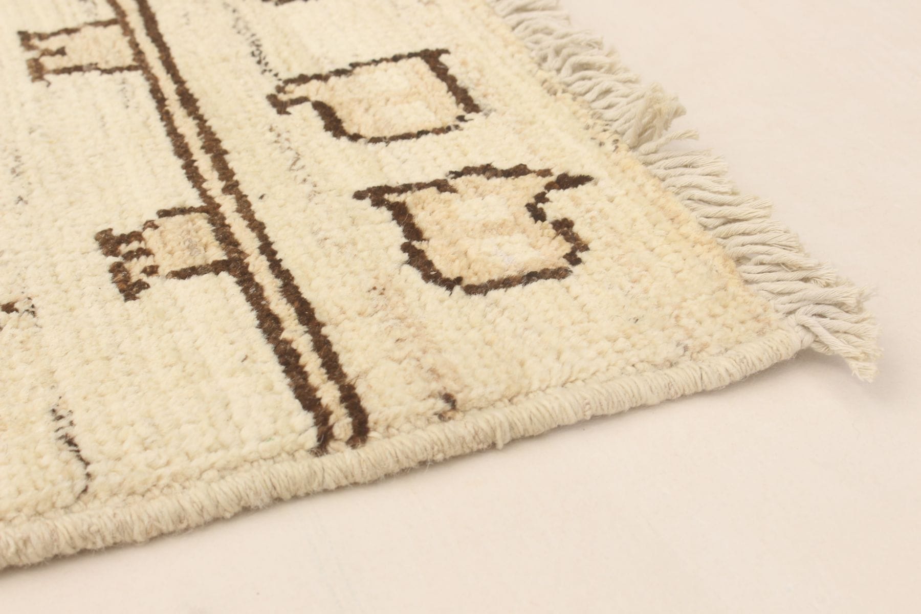 Close-up van een hoek van een beige tapijt met bruine geometrische patronen en franjeranden, gelegd op een licht tapijt.