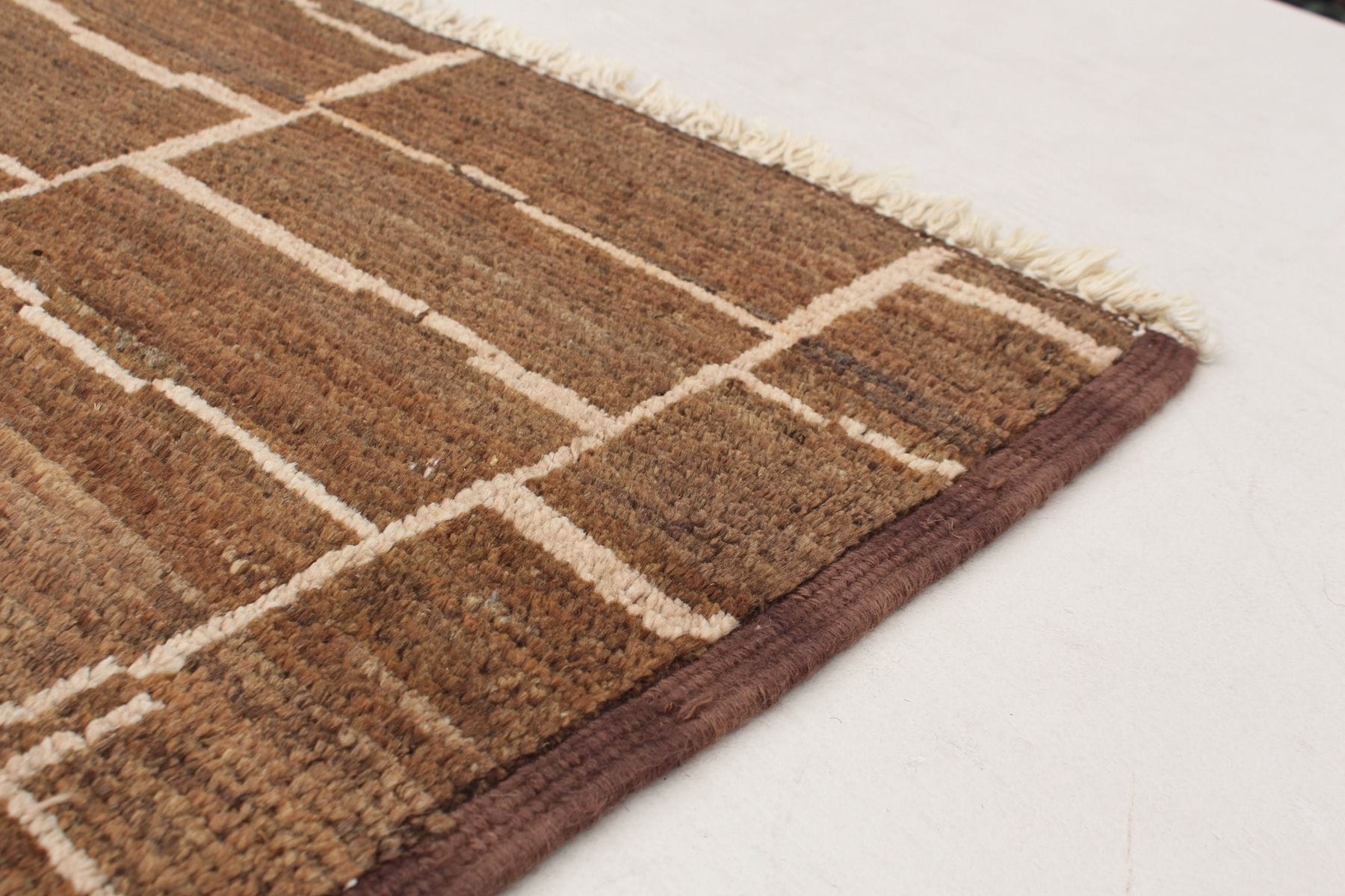 Een hoek van een bruin geweven tapijt met witte geometrische patronen en franjeranden op een licht oppervlak.