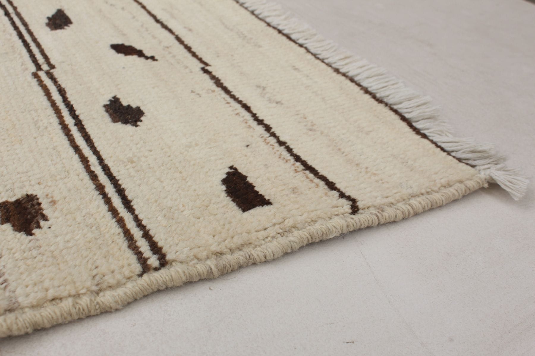 Close-up van een beige kleed met franjeranden, dunne bruine strepen en verspreide donkerbruine vlekken op een grijze vloer.