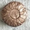 De MAROKKAANSE BRONZEN VOETENBANK VAN IMITATIELEER is een ronde, bronskleurige poef met een gestikt geometrisch patroon erop, prachtig geplaatst op een lichtgekleurd, gestructureerd vloerkleed.