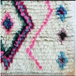 Een deel van een wollen vloerkleed met abstracte patronen in roze, blauw en groen op een witte achtergrond.