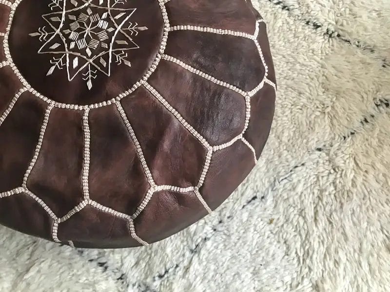 Ronde, bruine leren poef met ingewikkeld wit stiksel op een licht geblokt tapijt.