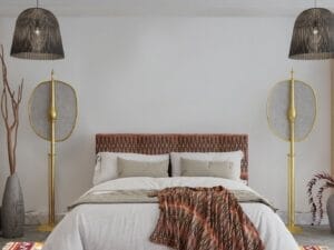 Een slaapkamer met een tweepersoonsbed, beige beddengoed, een hoofdeinde met patroon en deken. Aan beide zijden staan dubbele lampen. Het decor bestaat uit hoge rustieke vazen, een kleurrijk geometrisch vloerkleed en hangende rieten lampen.