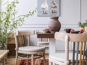 Een eethoek met twee stoelen, een ronde tafel, een vaas en serviesgoed. Aan de muur hangt een schilderij van twee ganzen. Aan de linkerkant staat een potplant en op de vloer ligt een tapijt met kleurrijke patronen.