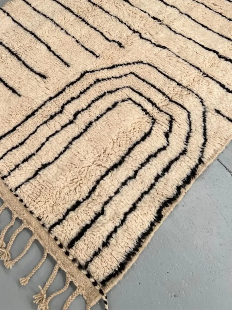 Een beige tapijt met zwarte geometrische lijnen en franje op een grijze vloer.