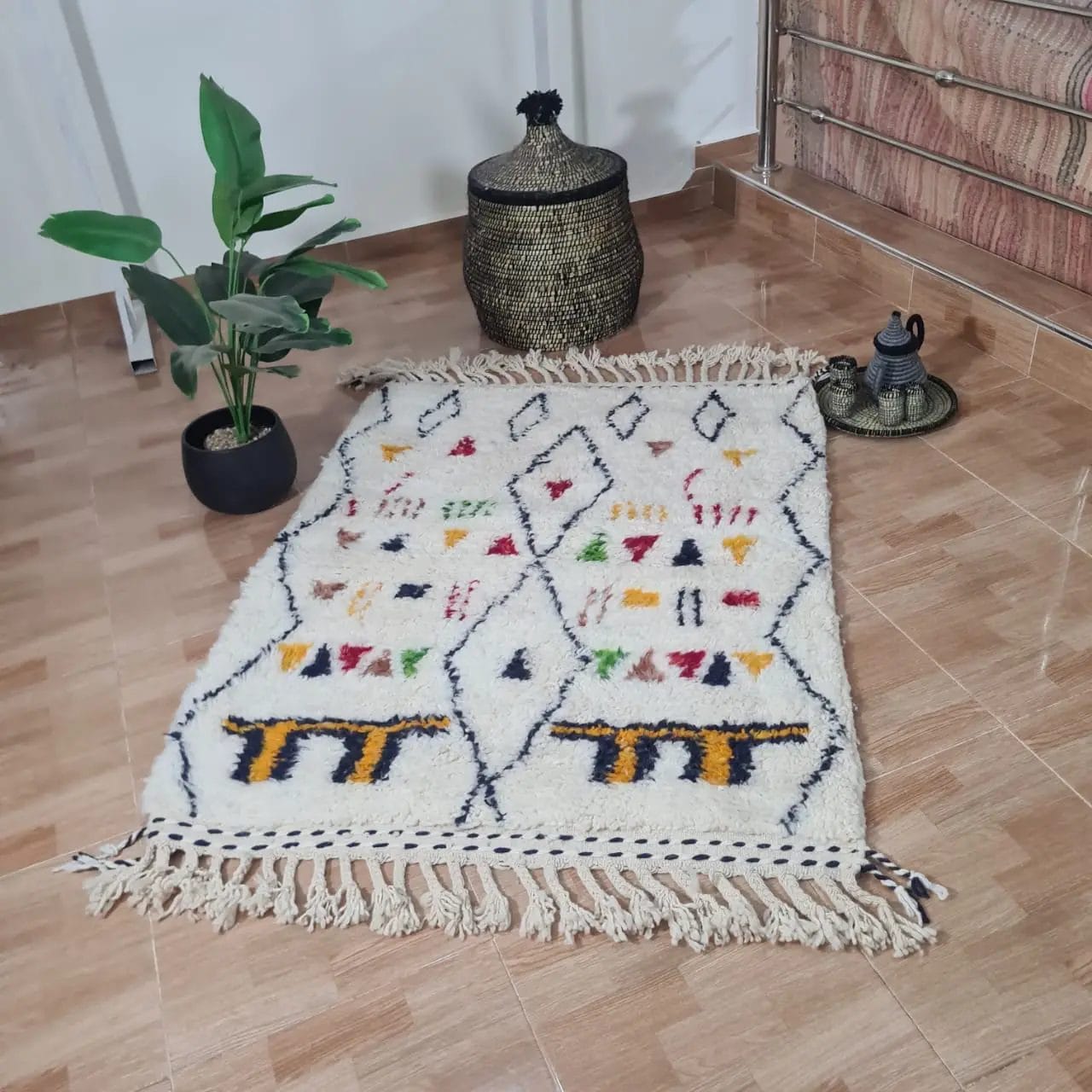 Het 100% HANDGEMAAKTE MAROKKAANSE TAPIJTEN TAPIJT 150-100 cm nr 42, met witte en veelkleurige geometrische patronen, ligt op een betegelde vloer naast een potplant, een gevlochten mand en een klein decoratief object.