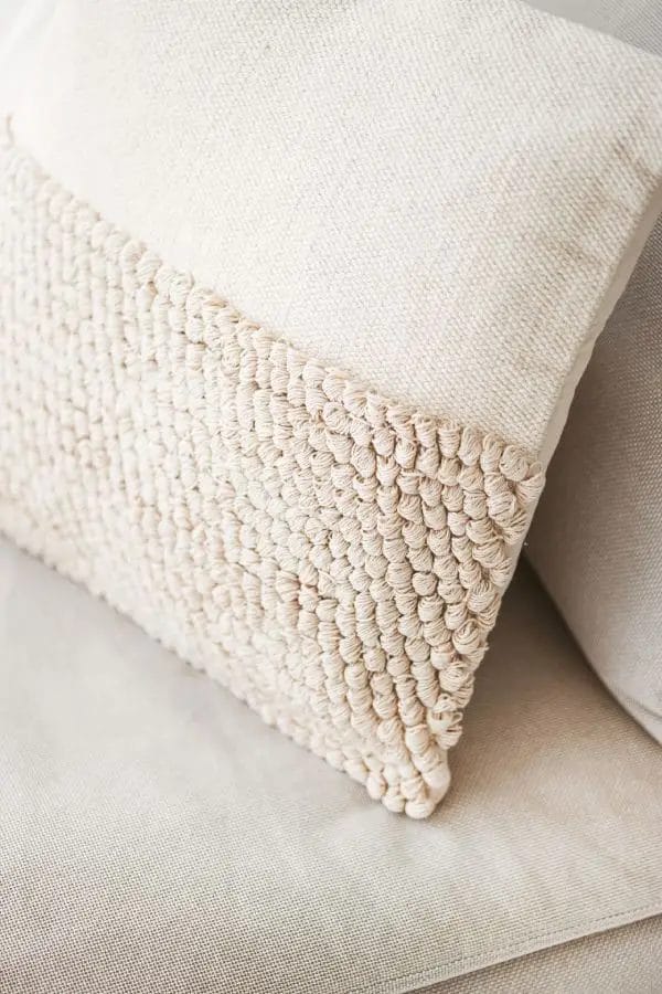 Een beige sierkussen met een geweven structuur aan de ene kant en rust op een lichtgekleurd stoffen oppervlak.