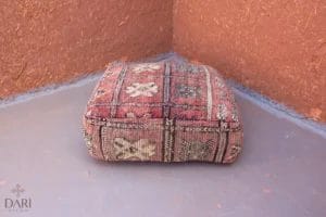 Een vierkant kussen met een patroon van bloemenpatronen ligt op een grijze vloer in de hoek van een kamer met bruine muren.