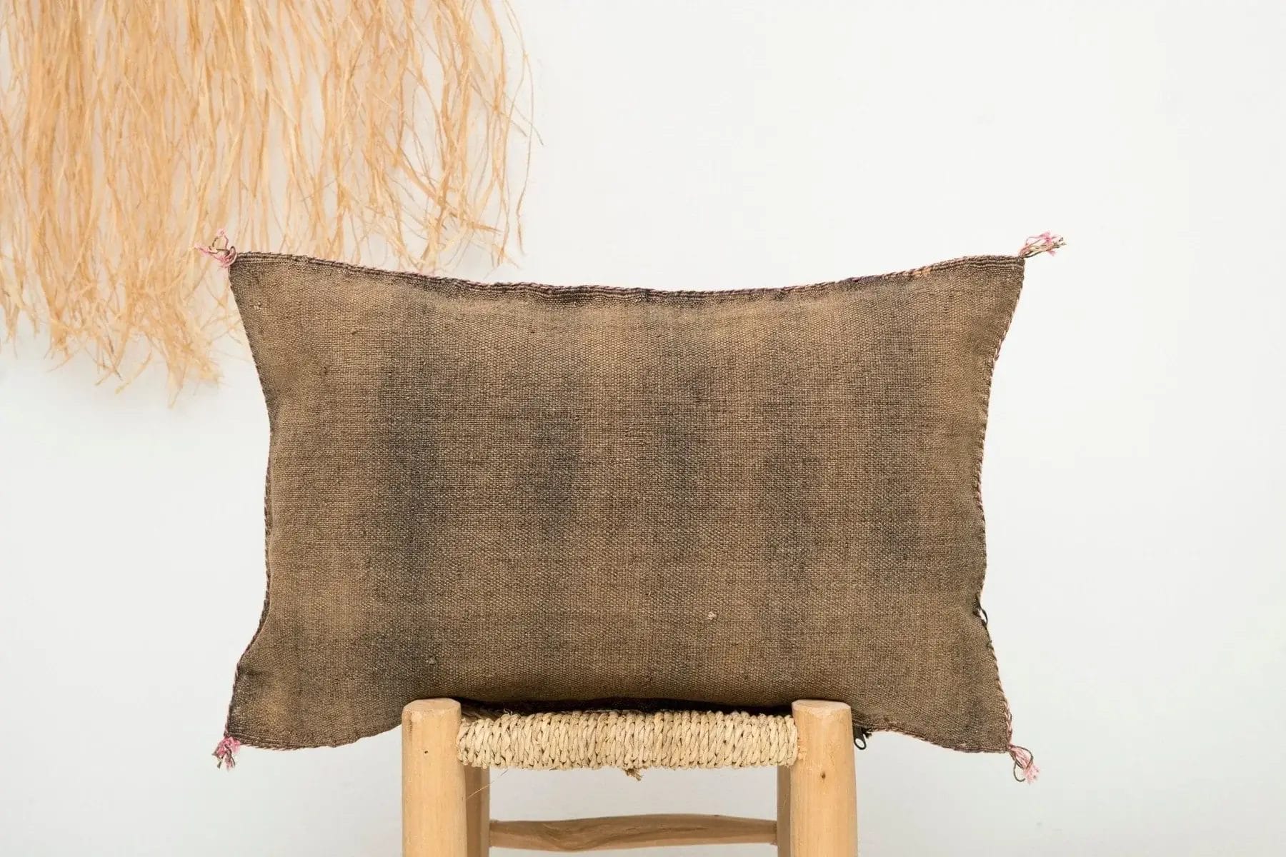 Een rechthoekig jute kussen met gerafelde randen rust op een gevlochten krukje, tegen een witte muur met strodecoratie.