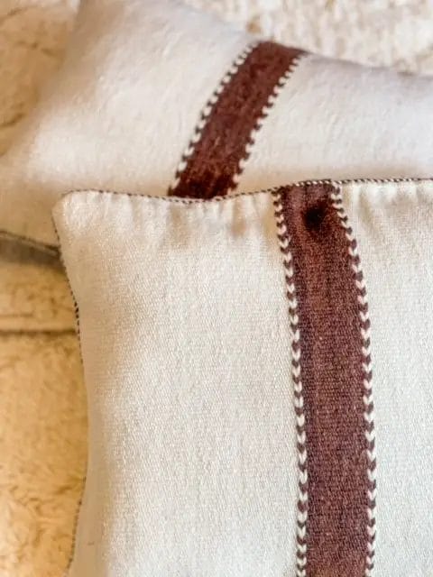 Twee beige kussens met bruine strepenpatronen en decoratieve stiksels liggen op een gestructureerd oppervlak.