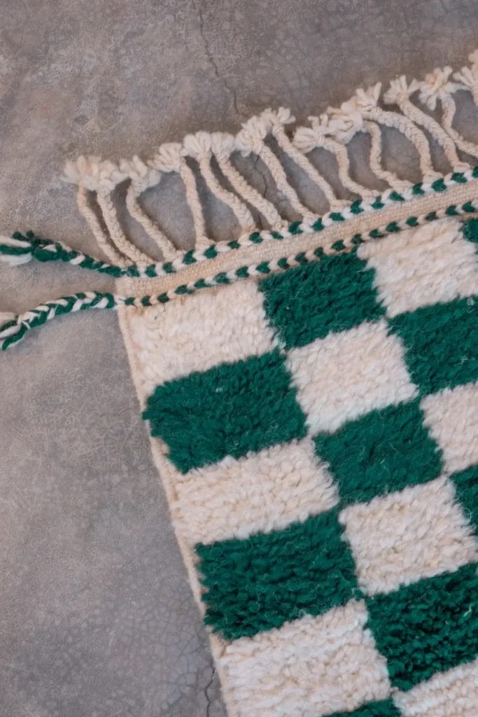 Een geruit vloerkleed met groene en witte vlakken, met franjes langs de rand, gelegd op een grijs gebarsten oppervlak.