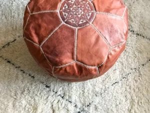 Een bruine leren poef met witte geborduurde patronen staat op een crèmekleurig vloerkleed met subtiele zwarte ruitvormen.